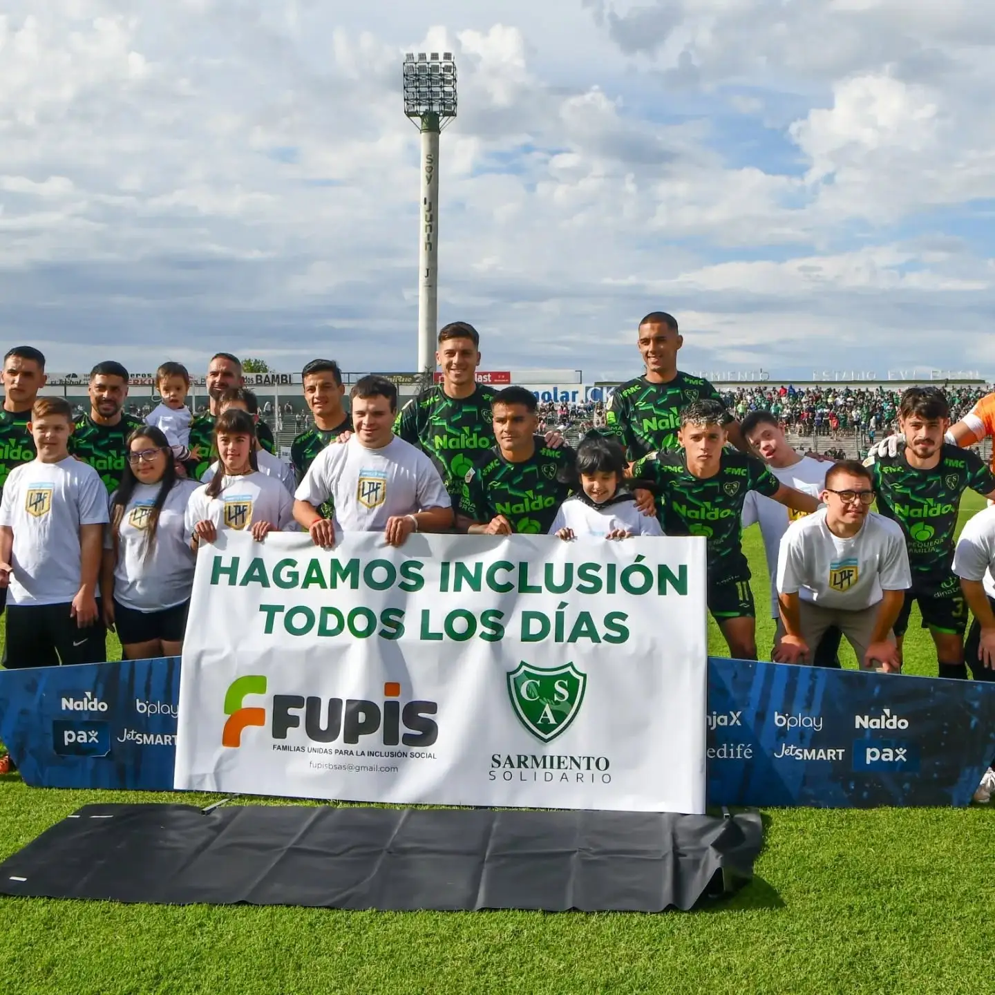 Chicas, chicos y jugadores de Sarmiento de Junín sostienen un banner que dice Hagamos inclusión todos los días junto al logo de FUPIS